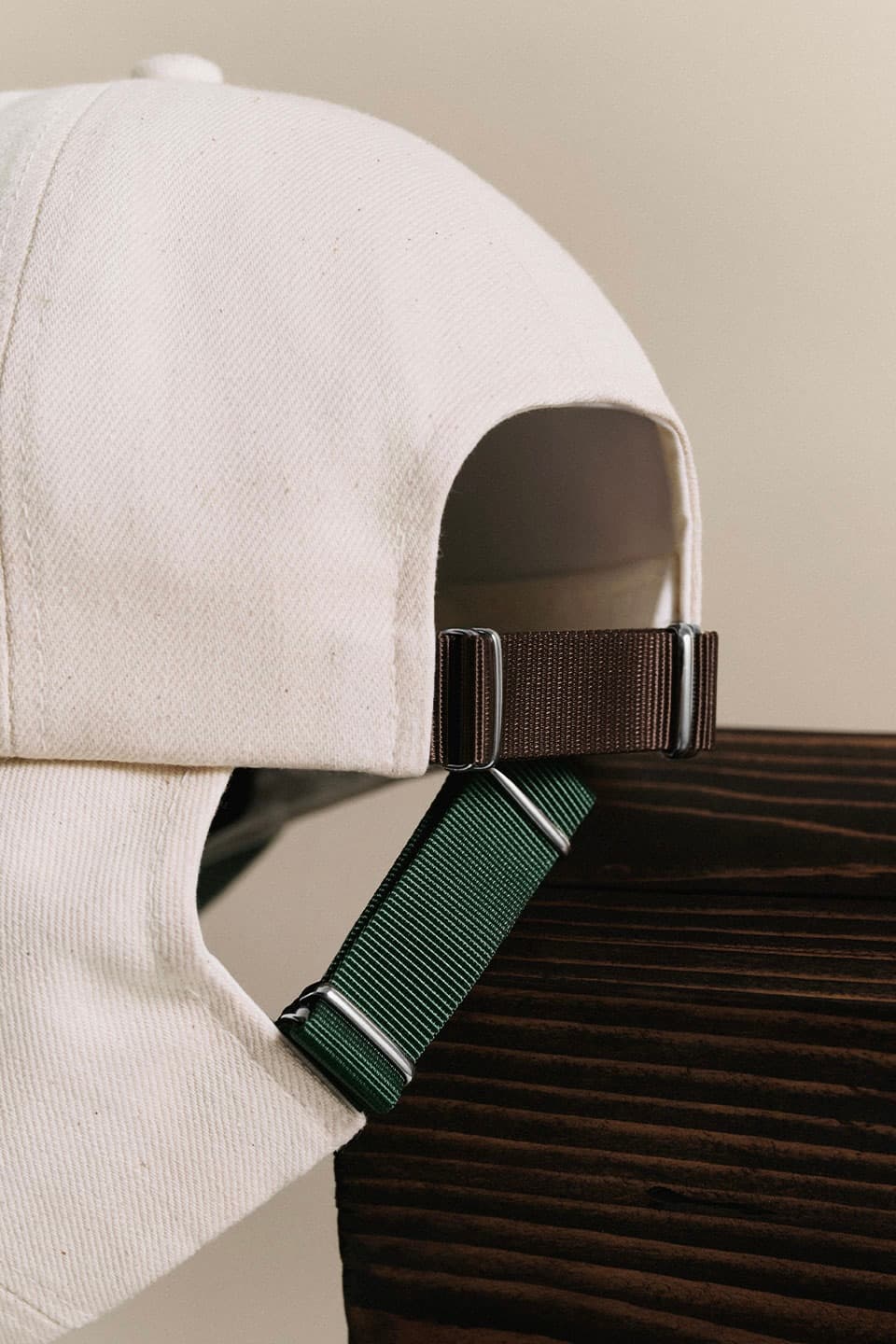 Close-up of a beige cap with brown and green adjustable straps resting on a dark wooden surface against a neutral background.