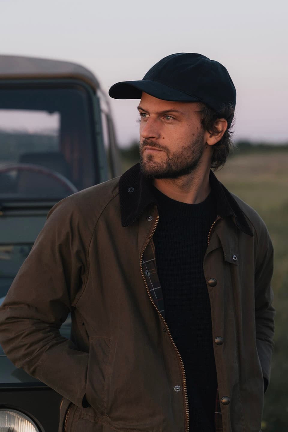Man with a beard and black cap stands by a rugged vehicle, wearing a brown jacket, looking into the distance with a contemplative expression.