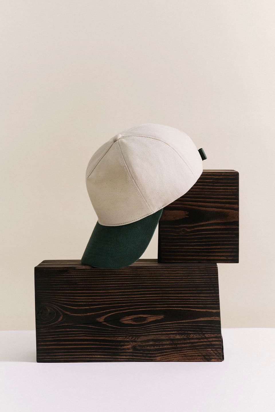 A beige baseball cap with a green brim is displayed on dark brown wooden blocks against a light beige backdrop.